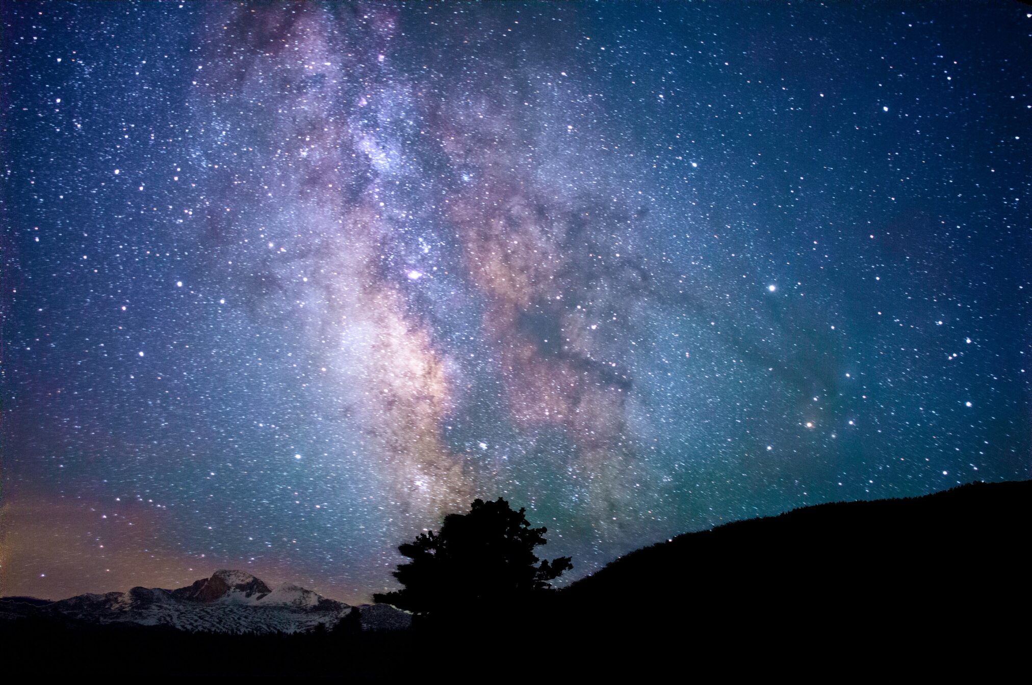 A night sky in shades of blues, purples, and pinks with a tree and mountains in the foreground.